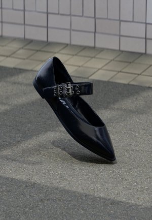 Černá špičatá bota na nízkém podpatku s širokým páskem se dvěma stříbrnými přezkami a kovovými průchodkami, umístěná vzpřímeně na asfaltu poblíž obložené zdi.