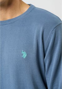 Blå strikket sweater med rund halsudskæring og ribbede manchetter; inkluderer et lille, broderet logo i teal på venstre bryst.