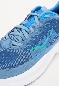 Baskets de sport bleues en maille et en matériau synthétique. Elles disposent d'un dessus texturé, de lacets bleus et d'un logo "HOKA" en blanc et vert.