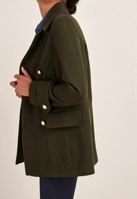 Olijfgroen wollen mix jas met een getailleerde pasvorm, dubbele knoopsluiting, gouden knopelementen en zijzakken, zij-aanzicht.
