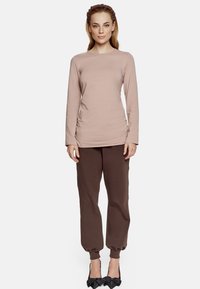 Jasno-beżowa bluzka o długim rękawie z okrągłym dekoltem i bocznym marszczeniem, w połączeniu z brązowymi zwężanymi spodniami dresowymi i czarnymi szpilkami.