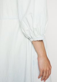 Abito azzurro chiaro con maniche a sbuffo, polsini arricciati e una texture liscia. Presenta un design semplice e una leggera lucentezza.