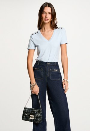 Kvinde iført en lyseblå V-hals skjorte med knappedetaljer og mørkeblå højtkaljede jeans, der holder en sort tekstureret håndtaske.