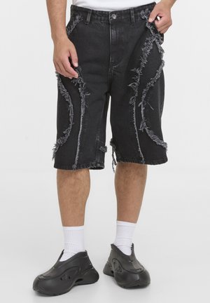 Sorte knælange denimshorts med flere lodrette flossete sømme, båret med hvide sokker og sorte slip-on sneakers.