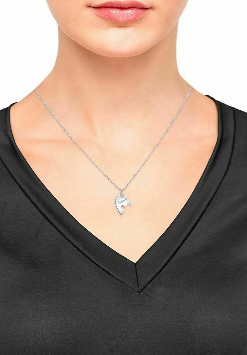 Silberne Halskette mit einem kleinen, geformten Anhänger, auf dem das Wort "Bär" steht, getragen von einer Person mit einem schwarzen V-Ausschnitt-Shirt, Nahaufnahme.