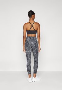 Fekete sportmelltartó keresztpántos hátpánthoz, magas derekú, szürke kamuflázs leggings-szel párosítva. A modell fehér sportcipőt visel, háttal áll.