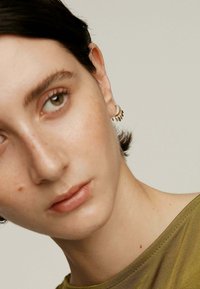 Acercamiento a una persona de piel clara, con cabello corto y oscuro, que lleva una blusa transparente de color verde oliva. Los pendientes de aro dorados presentan pequeños acentos en forma de hoja.