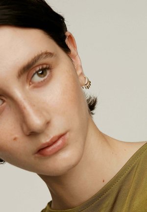 Acercamiento a una persona de piel clara, con cabello corto y oscuro, que lleva una blusa transparente de color verde oliva. Los pendientes de aro dorados presentan pequeños acentos en forma de hoja.