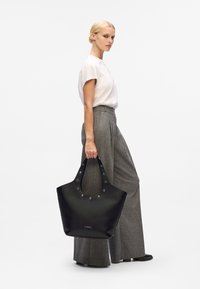 Sac tote en cuir noir avec des accents en métal argenté et de larges poignées, présentant un design spacieux et une texture lisse. Modèle portant un pantalon gris.