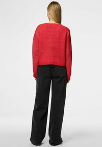 Röd stickad tröja med ribbat mönster, rund halsringning och kort skärning, kombinerad med vida svarta byxor. Bakifrån framhävs den lösa passformen.