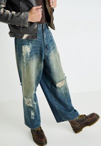 Vaqueros de mezclilla anchos con efecto desgastado, color azul desvaído, grandes rasgaduras y bajos deshilachados, combinados con botas marrones con cordones.