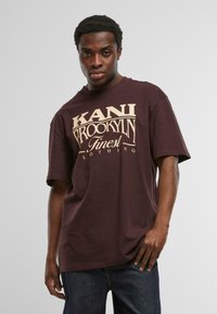 Bordeauxfarbene Baumwoll-T-Shirt mit großem cremefarbenem Schriftzug "KANI BROOKLYN Finest Clothing." Das Design ist schlicht mit kurzen Ärmeln.