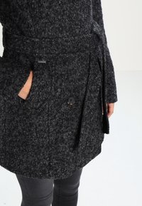 Abrigo de mezcla de lana negro con un acabado texturizado, que presenta un cinturón atado, un gran botón delantero y bolsillos laterales.