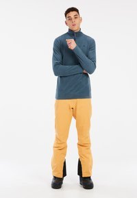 Maglione in pile blu con zip e colletto alto, abbinato a pantaloni impermeabili arancioni brillanti e stivali da neve neri. Design semplice, senza motivi visibili.