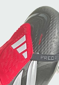 Zapato de fútbol rojo con parte superior texturizada en negro, acentos plateados y una superposición acolchada que presenta un logo y una correa etiquetada "PREDATOR."