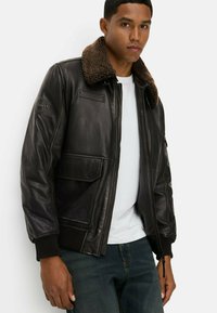 Schwarze Lederjacke mit einem braunen Kunstfellkragen, Reißverschluss vorne, gerippten Bündchen und zwei vorderen Taschen. Mit einem Logo-Patch auf der Schulter.