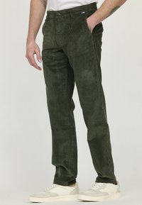 Pantalons en velours côtelé vert avec une coupe droite, dotés d'une fermeture à bouton et d'une texture subtile. Associés à des baskets blanches et beige.
