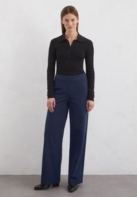 Cămașă polo neagră cu mâneci lungi, guler și nasturi, asociată cu pantaloni largi, cu talie înaltă, de culoare bleumarin și pantofi negri cu toc.