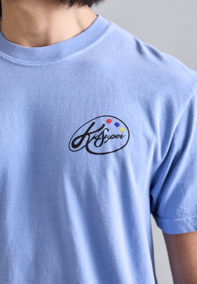 Light blue cotton t-shirt with a round black logo featuring the text "Krisuper" and colored dots in red, yellow, and blue. Short sleeves.