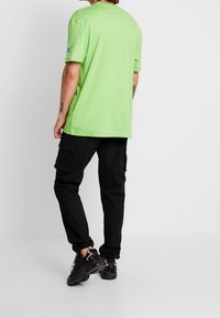 Green cotton t-shirt with short sleeves, round neck, and logo detail on the left sleeve, paired with black cargo pants and black sneakers.