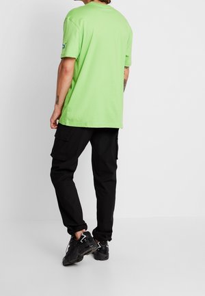 Grünes Baumwoll-T-Shirt mit kurzen Ärmeln, Rundhalsausschnitt und Logodetail auf dem linken Ärmel, kombiniert mit schwarzen Cargo-Hosen und schwarzen Sneakers.