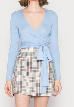 Vrouw draagt een lichtblauw, lang mouw wikkeltruitje, gestrikt in de taille, en een hoog geplaatste pastel geruite minirok.