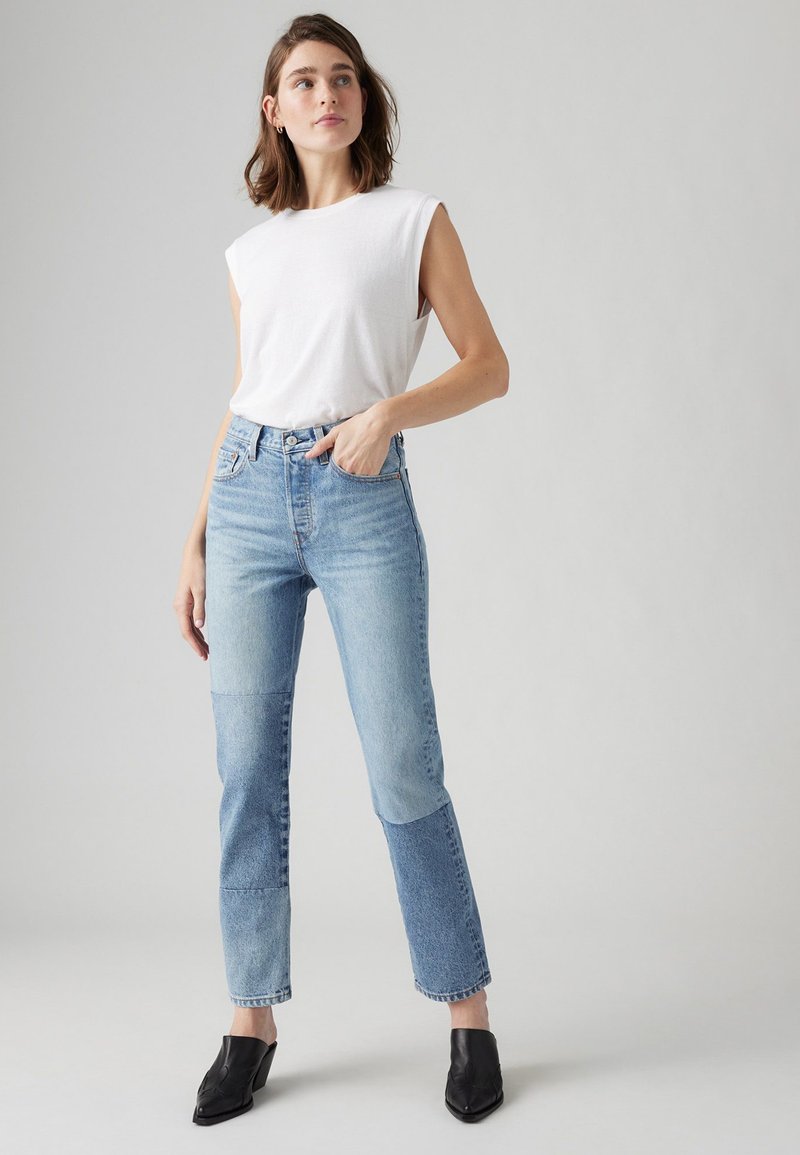 Top bianco senza maniche abbinato a jeans high-waisted azzurri chiari con design in patchwork e accenti blu scuro. Mules nere con tacco completano l'outfit.