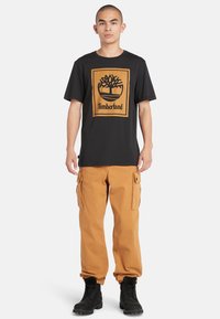 Zwarte katoenen T-shirt met een beige logo en boomgraphic. Gecombineerd met beige cargo pants met zijzakken en zwarte veterschoenen.
