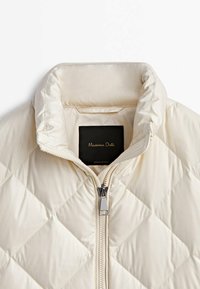 Veste matelassée couleur crème avec col montant et fermeture éclair centrale, dotée d'une étiquette noire à l'intérieur du col.