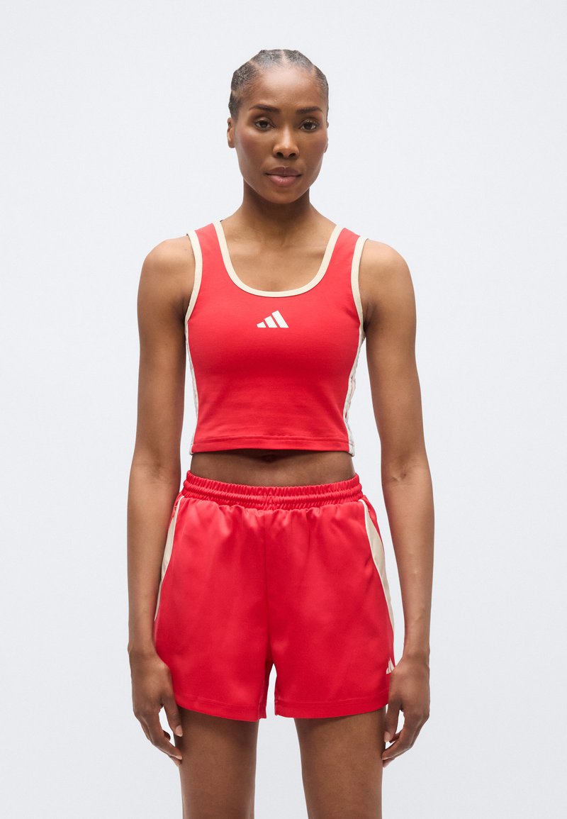 Κόκκινο αθλητικό crop top με λευκές λεπτομέρειες, που φέρει λογότυπο στο κέντρο. Συνδυάζεται με κόκκινα σορτς, και τα δύο από υλικό με λεία υφή.
