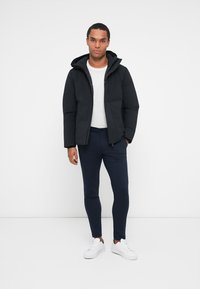 Černá péřová bunda s kapucí, dlouhými rukávy a předním zipem. Nositá přes bílý tričko a v kombinaci s námořnickými slim-fit kalhotami a bílými teniskami.