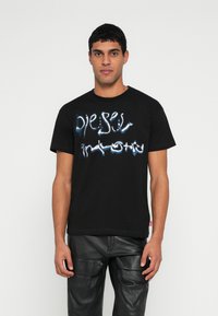 Μαύρο βαμβακερό μπλουζάκι με παραμορφωμένο λευκό και μπλε λογότυπο. Στρογγυλή λαιμόκοψη, κοντά μανίκια και κανονική εφαρμογή. Λεπτομέρεια υφής στο ύφασμα.