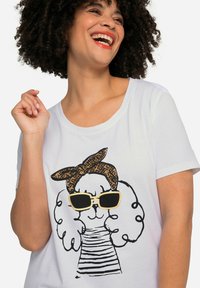 Hvid t-shirt med et tegneseriehundemotiv. Hunden har krøllet pels, solbriller og et mønstret pandebånd samt en stribet skjorte.