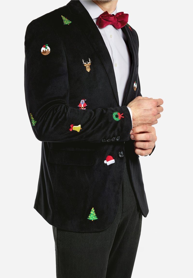 Man med svart kavaj med broderade julikoner, inklusive gran, krans, jultomte, julpudding, ren, klocka och röd fluga.