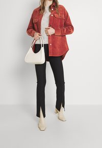 Chaqueta de camisa de pana roja, suéter gris de punto, pantalones negros con abertura en el dobladillo y botas beige con tacón. Bolso blanco con detalles de correas.