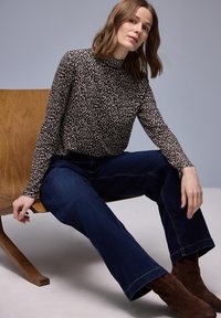 Haut à col roulé à motifs noir et blanc, jean évasé bleu foncé, et bottines marron. Modèle assise sur une chaise en bois sur un fond clair.