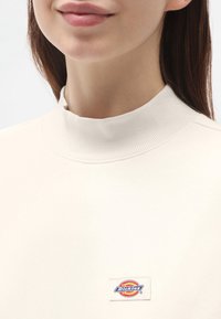 Sudadera de cuello alto color crema, hecha de material suave, con un pequeño parche de logo colorido de Dickies en el frente.