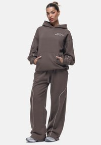 Bruine hoodie met een voorzak en witte letters; gecombineerd met bijpassende bruine sweatpants met contrasterende witte stiksels.