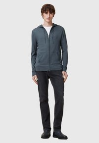 Felpa grigia con zip realizzata in tessuto morbido, con cappuccio regolabile, tasche frontali e polsini a coste, abbinata a jeans neri e scarpe nere.