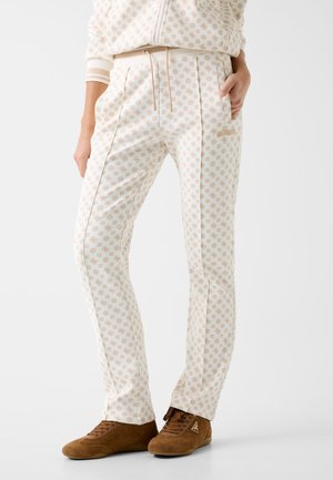 Osoba nosząca kremowe spodnie z jasnobrązowymi geometrycznymi wzorami, w połączeniu z dopasowaną kurtką i brązowymi zamszowymi trampkami.