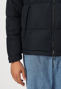 Sort pufferjakke med lynlås fortil, synlige stikninger og sidelommer, parret med lyseblå denim.