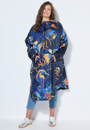 Kvinde iført en farverig regnfrakke med marint tema med søheste, fisk og koralmønstre, kombineret med korte jeans og hvide sneakers.