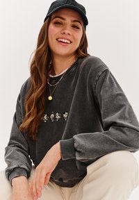 Šedá oversized mikina s pružnými manžetami, na přední straně s barevným obrázkem čtyř kreslených postaviček, v kombinaci s černou čepicí.