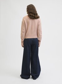 Γυναίκα με μαλλιά σε μήκος ώμου και κυματιστό στιλ, φοράει ένα ανοιχτό ροζ πουλόβερ και φαρδιά μπλε τζιν, στέκεται γυρισμένη προς τα πίσω.