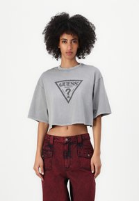 Grå avkortet T-skjorte med korte ermer som har en svart trekantet logo og spørsmålstegn. Kombinert med høyt liv røde denimbukser.