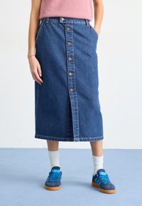 Fustă din denim de culoare albastru închis, de lungime medie, cu închidere frontală cu butoane și buzunare laterale. Purtați cu adidași albaștri și ciorapi albi până la glezne.