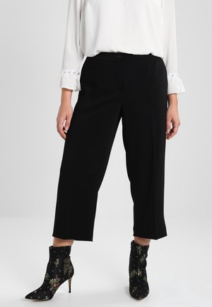 Femme portant un pantalon large court noir, un chemisier blanc avec des poignets à boutons perlés, et des bottines à talons hauts noires à motif floral.