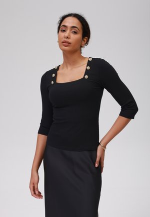 Black ribbed top with three-quarter sleeves, square neckline, and gold button accents on shoulders. Paired with a black skirt.