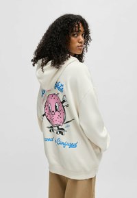 Un hoodie crème avec un grand motif de donut rose au dos, avec du texte bleu, fabriqué à partir d'un tissu doux, conçu pour le confort et le port décontracté.