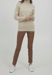 Frau in einem beigen strukturierten Rollkragenpullover, braunen Slim-Fit-Hosen und weißen Sneakers, stehend vor einem schlichten hellen Hintergrund.